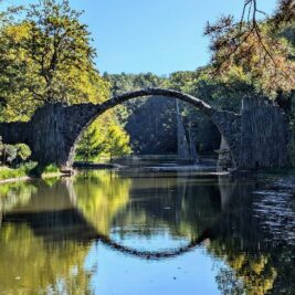 Rakotzbrücke in Gablenz, Kromlau umgeben von dichtem Herbstwald und dem ruhigen Rakotzsee, der das Bild der halbkreisförmigen Steinbrücke spiegelt.