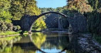 Rakotzbrücke in Gablenz, Kromlau umgeben von dichtem Herbstwald und dem ruhigen Rakotzsee, der das Bild der halbkreisförmigen Steinbrücke spiegelt.